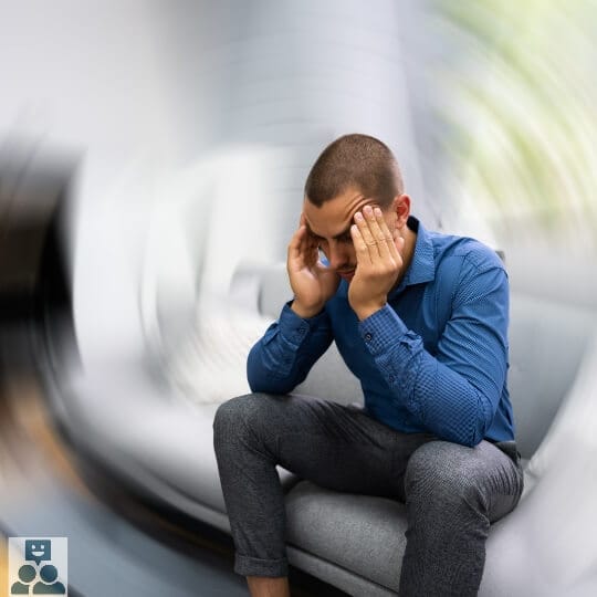 Beroertes, man zit op de bank op houdt zijn hoofd vast en heeft zichtbaar hoofdpijn. Man draagt een blauw overhemd en een grijze broek